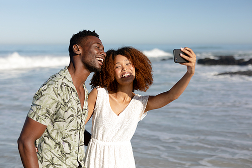 A mixed race couple enjoying free time on beach on a sunny day together, taking photos with a smartphone and holding each other with sun shining on their faces.