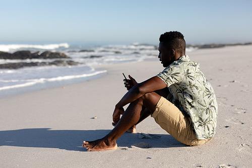 An attractive African American man enjoying free time on beach on a sunny day, sitting on sand, using his smartphone.