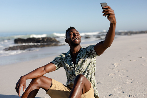 An attractive African American man enjoying free time on beach on a sunny day, sitting on sand, using his smartphone, taking photos and smiling.