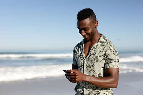 An attractive African American man enjoying free time on beach on a sunny day, standing on sand, using his smartphone and smiling with sun shining on his face.