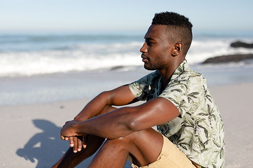 An attractive African American man enjoying free time on beach on a sunny day, wearing a Hawaiian shirt, sitting on sand and admiring the view with sun shining on his face.