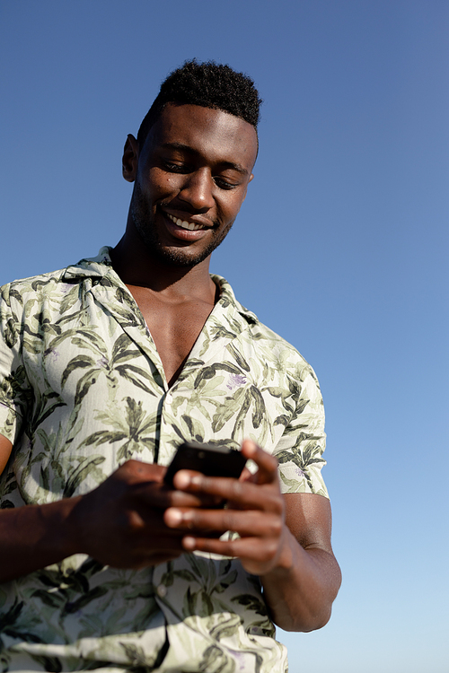 An attractive African American man enjoying free time on beach on a sunny day, standing, using his smartphone and smiling.
