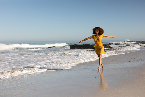 A happy, attractive mixed race woman with her arms outstretched enjoying free time on beach on a sunny day, wearing a yellow dress, walking with sun shining on her face.