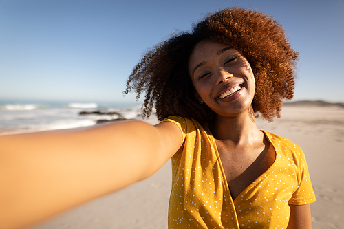 A portrait of a happy, attractive mixed race woman enjoying free time on beach on a sunny day, wearing a yellow dress, standing on sand, taking selfie with sun shining on her face.