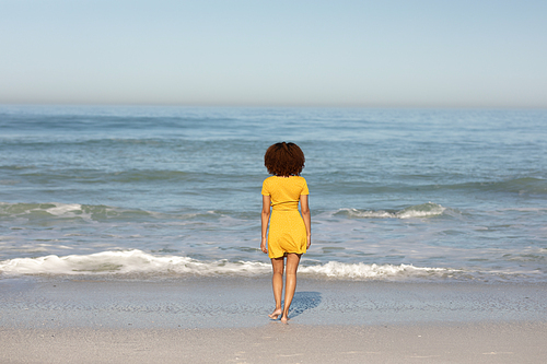 A rear view of a happy, attractive mixed race woman enjoying free time on beach on a sunny day, wearing a yellow dress, standing on sand, facing the sea with sun shining behind her.