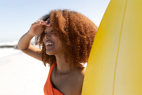 A happy, attractive mixed race woman enjoying free time on beach on a sunny day, smiling, having fun, standing with her surfboard, sun shining on her face.