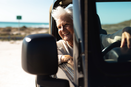 Front view of a senior Caucasian woman at the beach in the sun, sitting behind the wheel in the driving seat of a car, looking out of the side window and smiling