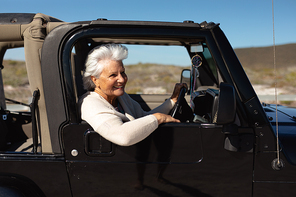 Side view of a senior Caucasian woman at the beach in the sun, sitting behind the wheel in the driving seat of a car, looking out of the side window and smiling