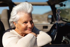 Side view close up of a senior Caucasian woman at the beach in the sun, sitting in the driving seat of a car, leaning and looking out of the side window and smiling