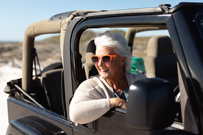 Side view of a senior Caucasian woman at the beach in the sun, sitting behind the wheel in the driving seat of a car, wearing sunglasses and looking out of the side window smiling