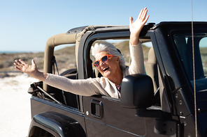 Front view of a senior Caucasian woman at the beach in the sun, sitting in the driving seat of a car, wearing sunglasses and looking out of the side window, raising her hands and smiling