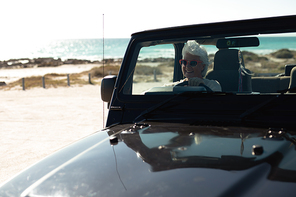 Front view of a senior Caucasian woman sitting behind the wheel in the driving seat of a car at the beach, wearing sunglasses and smiling, seen through the windscreen