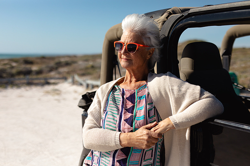 Front view of a senior Caucasian woman at the beach in the sun, leaning against a car, wearing sunglasses and looking away smiling