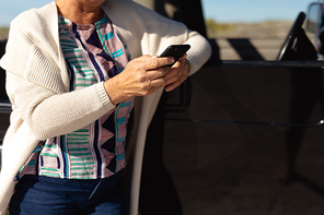 Side view mid section of a senior Caucasian woman at the beach in the sun, leaning against a car and using her smartphone