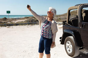 Side view of a senior Caucasian woman at the beach in the sun wearing sunglasses, standing beside her car and using a smartphone to take a selfie