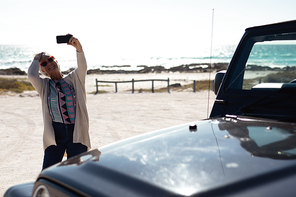 Front view of a senior Caucasian woman at the beach in the sun wearing sunglasses, standing beside her car and using a smartphone to take a selfie