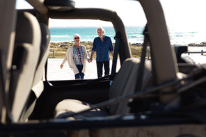 Front view of a senior Caucasian couple at the beach in the sun, holding hands and walking towards the camera, seen through a car in the foreground