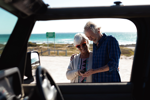Front view of a senior Caucasian couple at the beach in the sun, embracing, smiling and using a smartphone together, seen through the side window of a car in the foreground