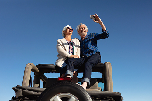 Low angle front view of a senior Caucasian couple sitting on the roof of their car at the beach in the sun, embracing, smiling and using a smartphone to take a selfie