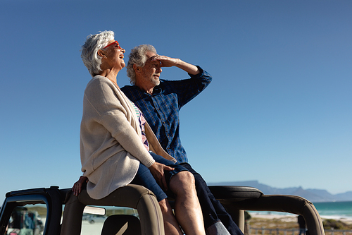 Side view of a senior Caucasian couple sitting on the roof of their car at the beach in the sun, admiring the view and smiling