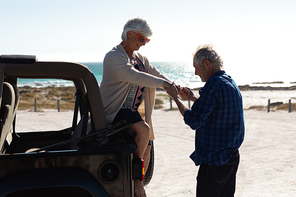 Side view of a senior Caucasian couple at the beach in the sun, the man helping the woman out of their car, holding her hand and smiling