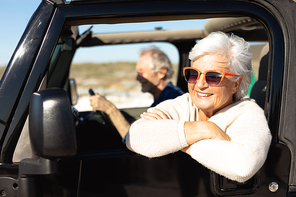 Side view of a senior Caucasian couple sitting in their car, driving to the beach in the sun, the man at the wheel and the woman in the foreground, wearing sunglasses and leaning out of the passenger window, focus on the foreground