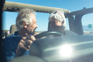 Front view of a senior Caucasian couple sitting in their car, driving to the beach in the sun, the man at the wheel, looking at each other and smiling, seen through the windscreen, lens flare