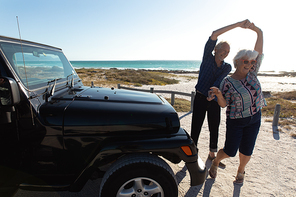 Front view of a senior Caucasian couple at the beach in the sun, holding hands and dancing beside their car, smiling and having fun