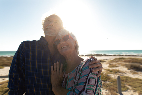 Front view close up of a senior Caucasian couple at the beach in the sun, standing, smiling and embracing, with the sea in the background, backlit