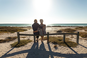 Rear view of a senior Caucasian couple at the beach in the sun, sitting on a fence, admiring the sea view and embracing