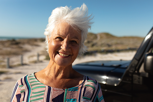 Portrait close up of a senior Caucasian woman at the beach in the sun, looking to camera and smiling beside her car