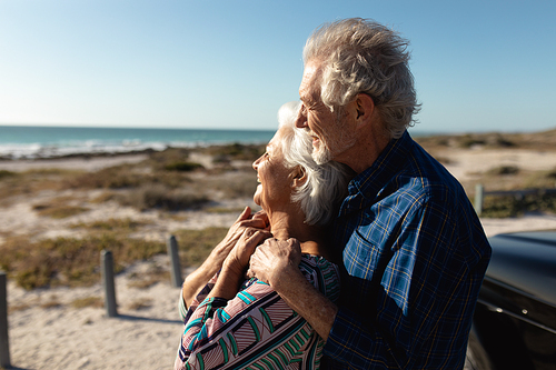 Side view of a senior Caucasian couple at the beach in the sun, embracing, leaning against their car and enjoying the sea view