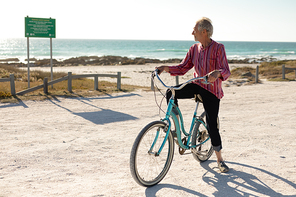 Front view of a senior Caucasian man at the beach in the sun, sitting on a bicycle and looking away, the sea in the background