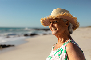 Portrait of a senior Caucasian woman at the beach in the sun, wearing a sun hat, looking out to sea and smiling, with blue sky in the background
