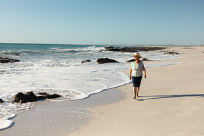 Front view of a senior Caucasian woman at the beach in the sun, wearing a sun hat and walking barefoot in the sand and smiling, with sea and blue sky in the background