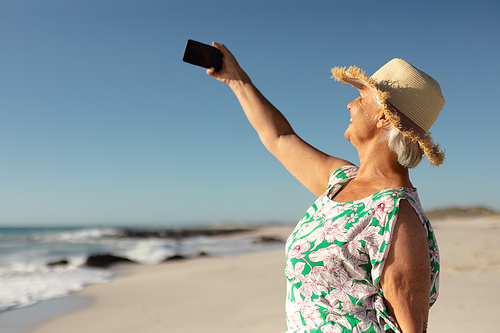 Side view of a senior Caucasian woman at the beach in the sun, wearing a sun hat, holding a smartphone, taking a selfie and smiling, with sea and blue sky in the background