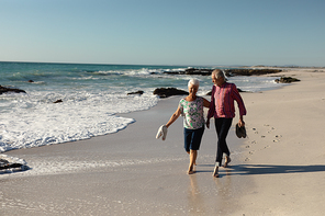 Front view of a senior Caucasian couple at the beach in the sun, holding their shoes and walking barefoot, smiling and talking, with sea and blue sky in the background