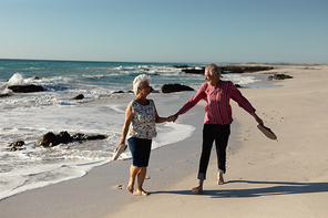 Front view of a senior Caucasian couple at the beach in the sun, holding their shoes and walking barefoot holding hands, smiling and talking, with sea and blue sky in the background