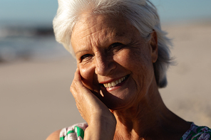 Portrait close up of a senior Caucasian woman at the beach in the sun, looking to camera, touching her face and smiling