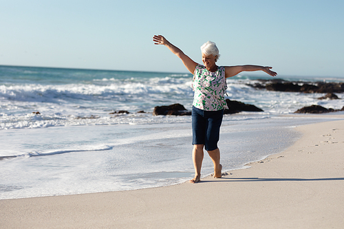 Front view of a senior Caucasian woman at the beach in the sun, walking with arms outstretched and smiling, with blue sky and sea in the background