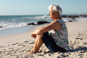 Side view of a senior Caucasian woman at the beach in the sun, sitting on the sand and looking out to sea, with blue sky in the background