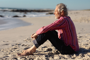 Side view of a senior Caucasian man at the beach in the sun, sitting on the sand, with blue sky in the background
