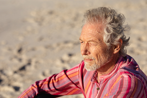 Side view close up of a senior Caucasian man at the beach in the sun, sitting on the sand, smiling and looking out to sea