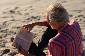 High angle view of a senior Caucasian man at the beach in the sun, sitting on the sand and reading a book