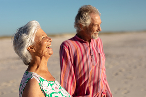 Side view of a senior Caucasian couple at the beach in the sun, holding hands and looking out to sea, the woman laughing, with blue sky in the background