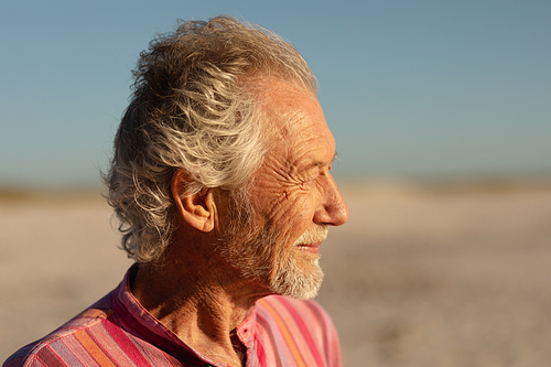 Side view close up of a senior Caucasian man at the beach in the sun, looking away and smiling, with blue sky in the background