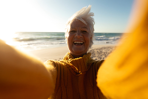 Front view of a senior Caucasian woman at the beach in the sun, smiling to camera and reaching out taking a selfie with a device out of shot