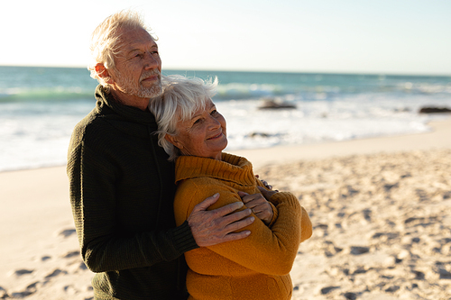 Side view of a senior Caucasian couple at the beach wearing sweaters, embracing and smiling, with blue sky in the background