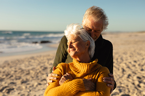 Front view of a senior Caucasian couple at the beach wearing sweaters, embracing and smiling, with blue sky and sea in the background