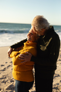 Side view close up of a senior Caucasian couple at the beach wearing sweaters, embracing and smiling, with blue sky and sea in the background